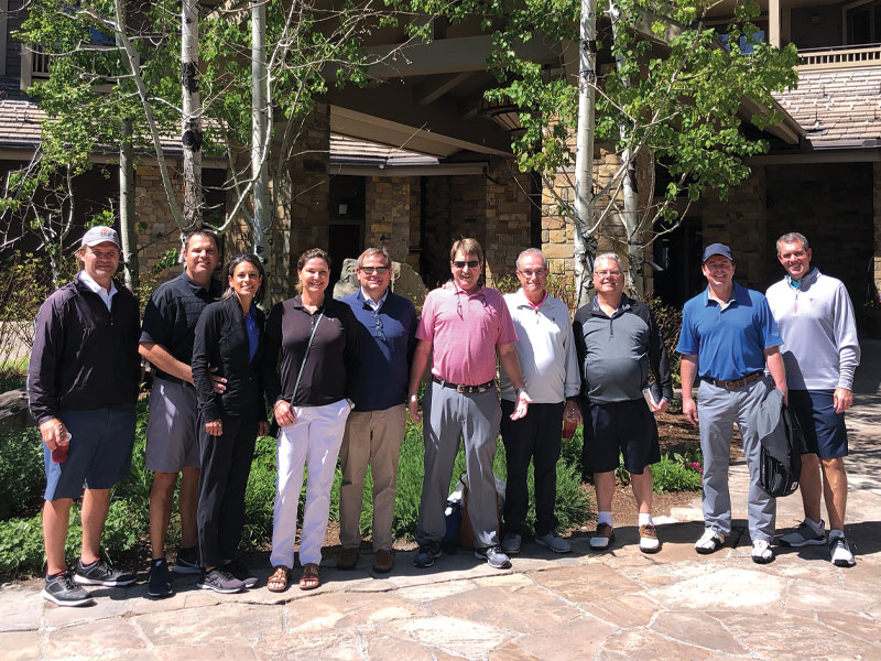 Eleven golfers standing in front of a stone clubhouse on a sunny day.